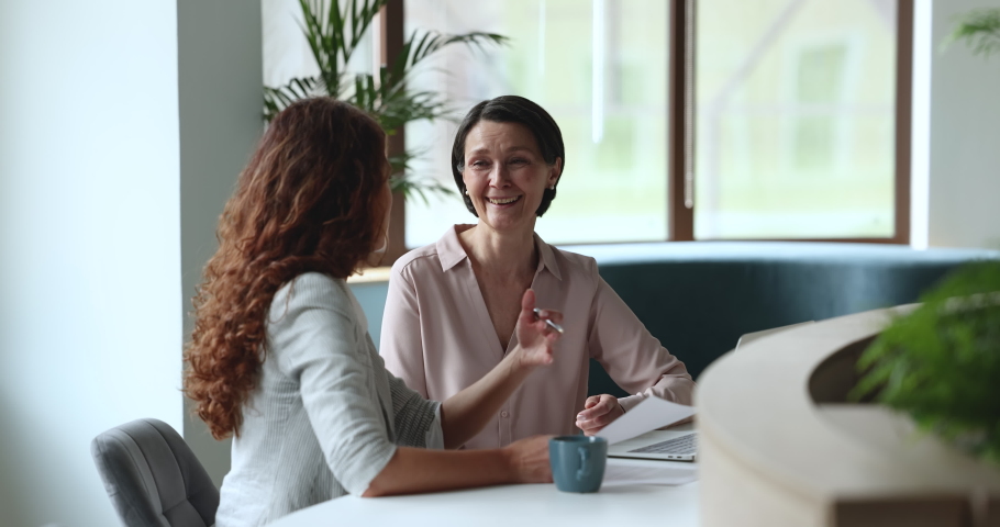 Two attractive businesswomen, middle-aged, millennial client and sales manager sit at desk discuss contract details, discuss agreement or paperwork meet in modern office. Negotiations, deal, commerce Royalty-Free Stock Footage #1097249881