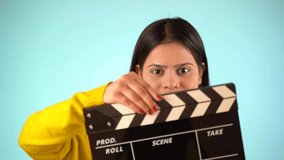 Beautiful young Asian Indian woman standing holding clapperboard, clapper board used in film making, isolated on blue color background studio portrait