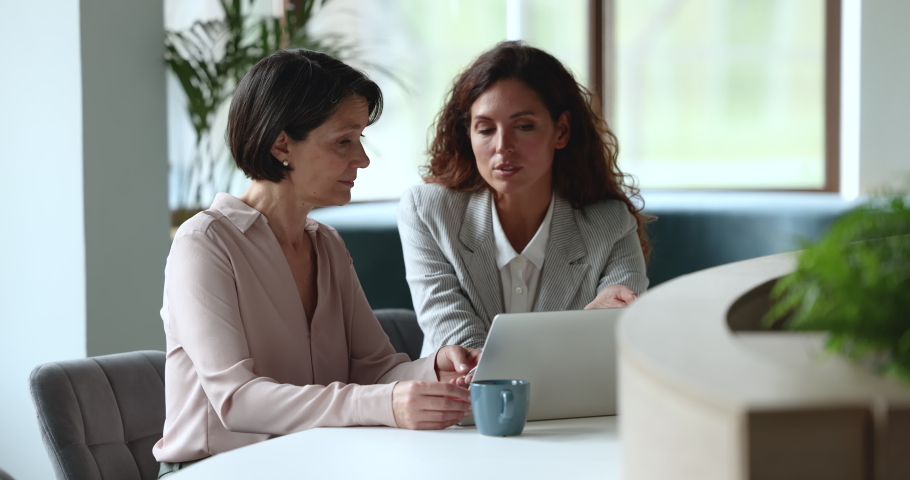 Hispanic millennial woman and middle-aged female colleagues working together meet in office staring at laptop screen, learn new software, younger mate helps to older with business app use modern tech Royalty-Free Stock Footage #1097251357
