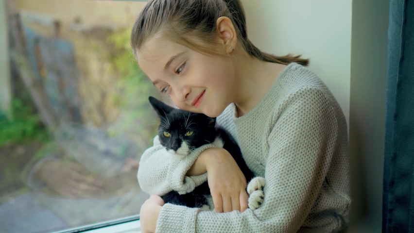 Happy girl sits on windowsill and looks out window with his cat black and white pet. Save lifes, no war, world peace, stop conflict, born love. Childhood in difficult times | Shutterstock HD Video #1097253411