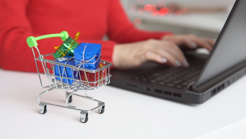Shopping cart with colorful gifts on table. Woman using laptop on the background. Shopping online during holidays. | Shutterstock HD Video #1097253415