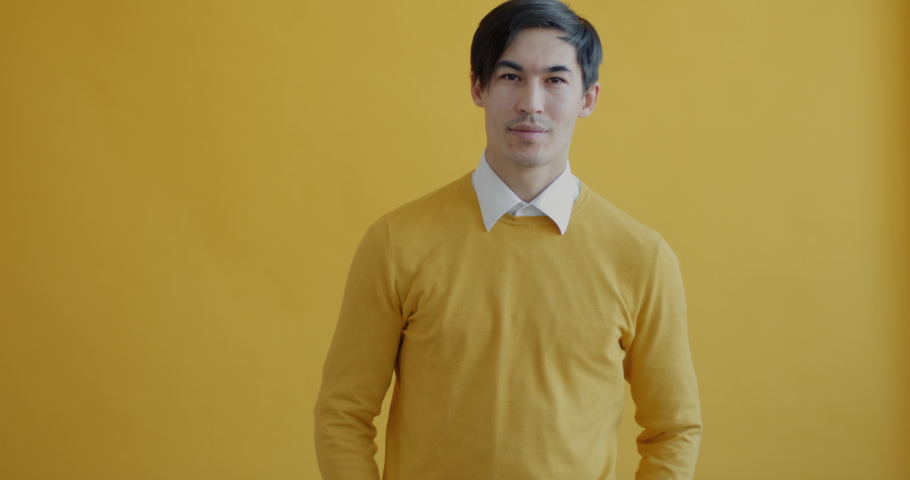 Portrait of friendly Asian man waving hands and smiling looking at camera standing on yellow background. Peope and positive emotions concept. | Shutterstock HD Video #1097254381