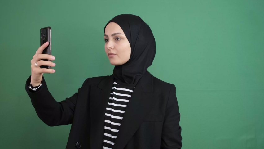 Muslim girl looking at herself through the phone camera, girl takes selfie in front of green screen, young girl tidy up the scarf | Shutterstock HD Video #1097255357