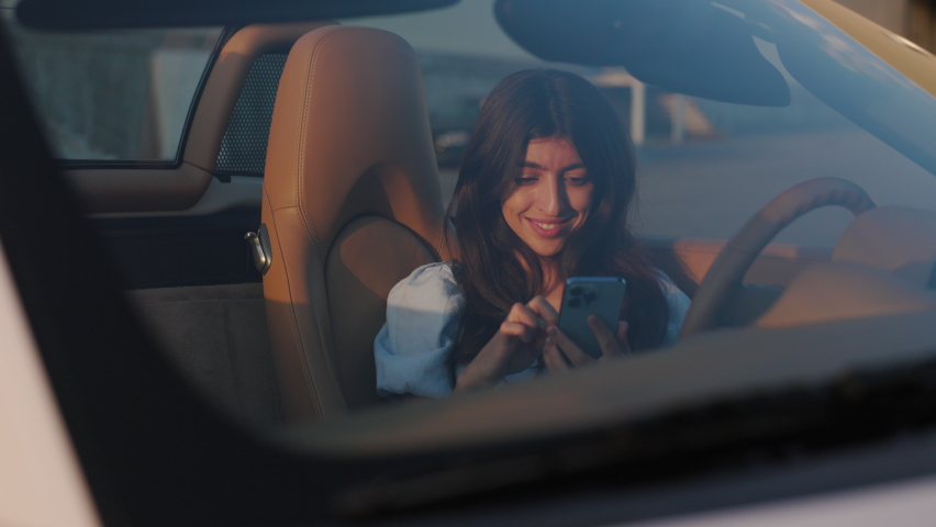 Beautiful stylish woman sits in her luxury white convertible and uses smartphone texting someone. View through the windshield. Sunset. | Shutterstock HD Video #1097256049