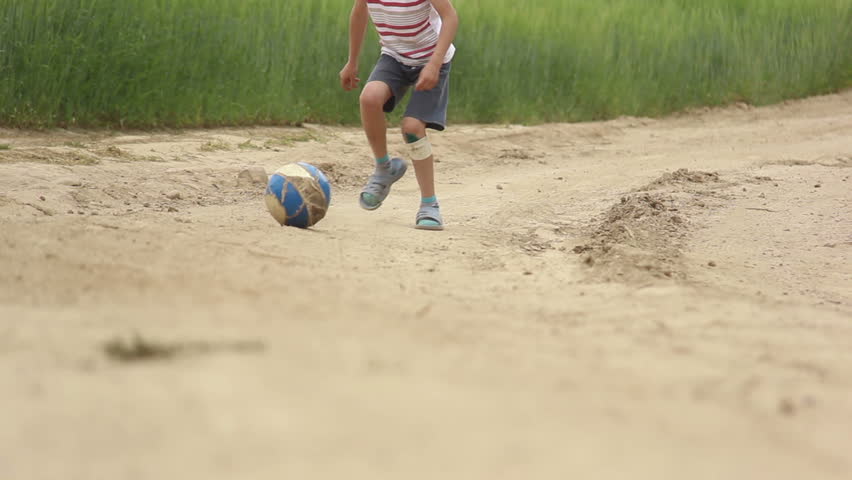 boy foot the ball on the sandy road, dust, boy playing with a ball on the sandy road near a wheat field, a boy with broken knees playing with a ball