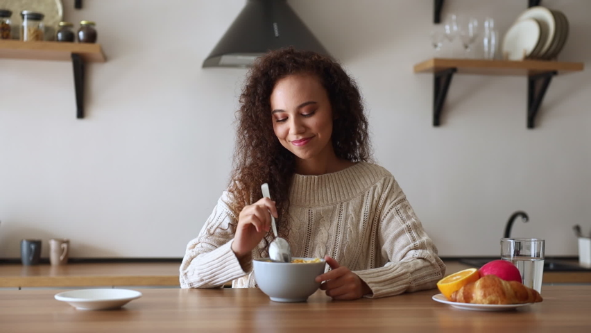 Young minded fun housewife woman of African American ethnicity in casual clothes look aside eat breakfast muesli cereals with milk fruit in bowl in light kitchen at home alone. Cooking food concept | Shutterstock HD Video #1097262797