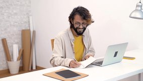Happy young creative entrepreneur laughing smiling writes in a notebook next to laptop in a modern workspace office at home. Freelance guy starting small business. High quality 4K video footage