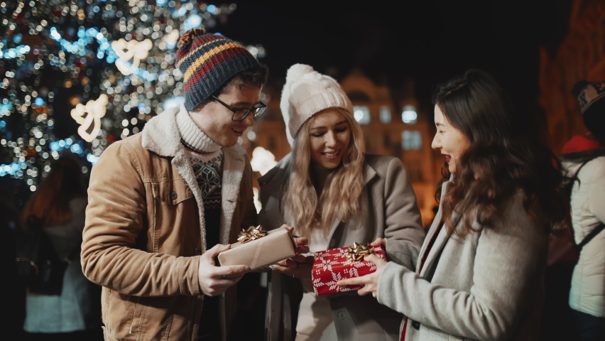 Happy multiracial friends present wrapped gifts each other at Christmas eve. People celebrate New year at decorated background outdoors at night party smile and hug. Winter holiday or vacation concept Royalty-Free Stock Footage #1097267649