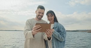 Young cute woman and beautiful elegant man using tablet video calling talking online standing on beach near a beautiful big lake enjoying trip. Lovely happy couple communicating smiling.