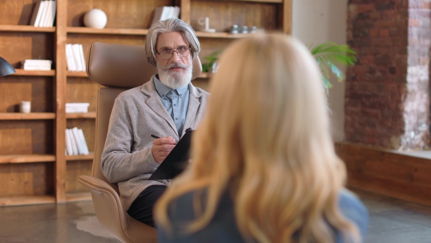 Elderly mature serious grey-haired bearded man psychologist having consulting session with patient. Young blonde client in psychotherapist's office. Handsome male giving advice solving mental problems | Shutterstock HD Video #1097267915
