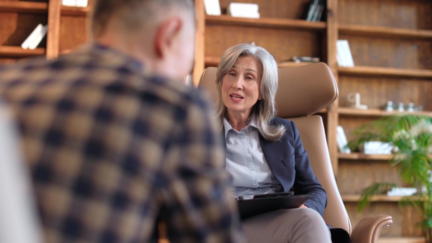 Over the shoulder view of mature professional psychologist woman helping consulting young depressed man having mental problems. Grey-haired attractive older female psychiatrist talking giving advice. | Shutterstock HD Video #1097267917