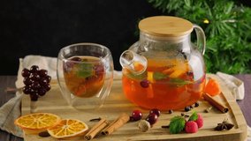 Black tea with fruit and herbs, cinnamon, red berries, woman in warm sweater takes a glass cup with hot tea from wooden table with transparent glass teapot, Christmas decorations, 4k video footage