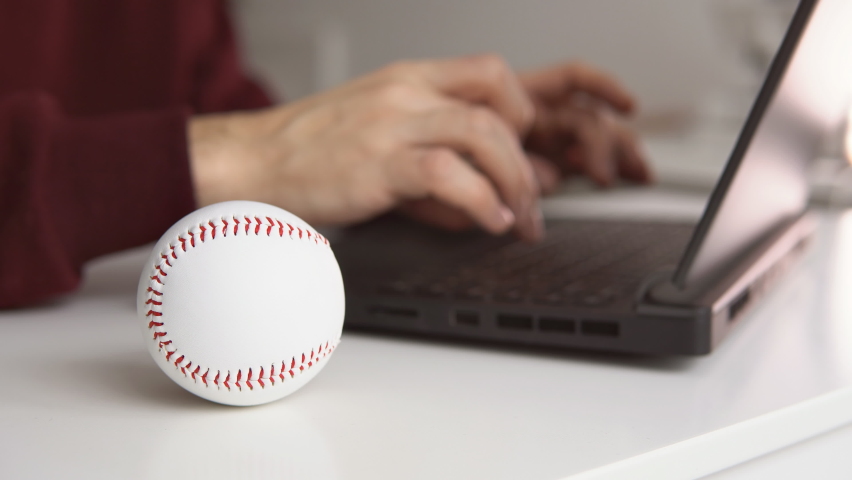 Young man checking baseball stats or writing baseball article on laptop. White leather ball on the table. Betting, gambling concept. | Shutterstock HD Video #1097269047