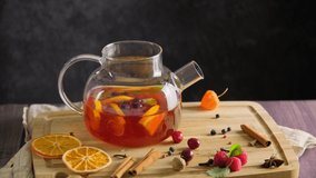 Hot tea with raspberry, orange fruit and herbs brewing in transparent glass teapot placed on wooden table on black background, stirring hot winter drink, slow motion 4k video footage