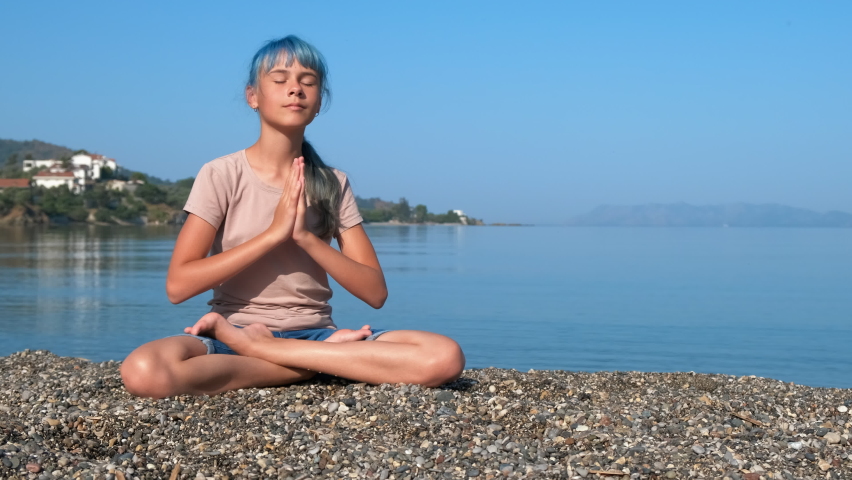 Thoughtfully namaste pose on sea. A view of beautiful young girl sitting in namaste thoughtful pose on the pebbles shore under sun. | Shutterstock HD Video #1097271313