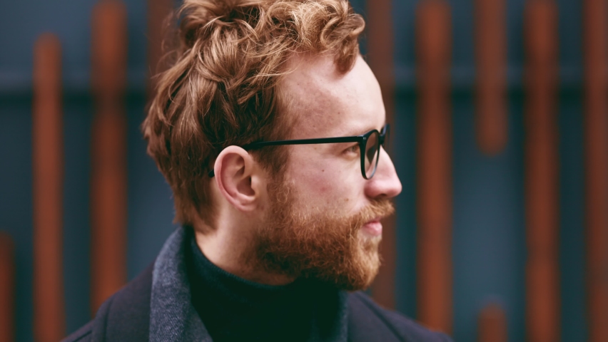 A young red-haired guy, a businessman in glasses, looks at the camera against the background of an urban style. Close up portrait Royalty-Free Stock Footage #1097273619
