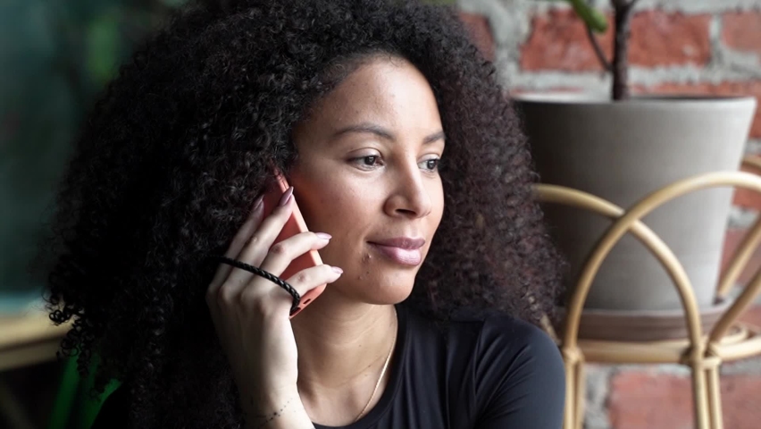 Black happy woman with African curls speaking on cellphone at home or cafe indoors. Communication with friends of relatives. Comfort relationships. Freelance | Shutterstock HD Video #1097278379