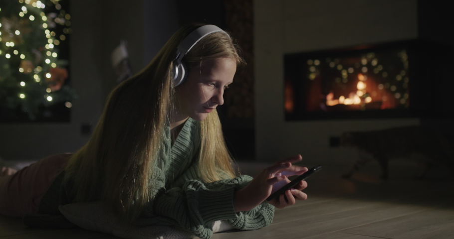 Child in headphones uses a smartphone. Lies on the floor in a room where a fireplace is burning and there is a large Christmas tree. | Shutterstock HD Video #1097278549