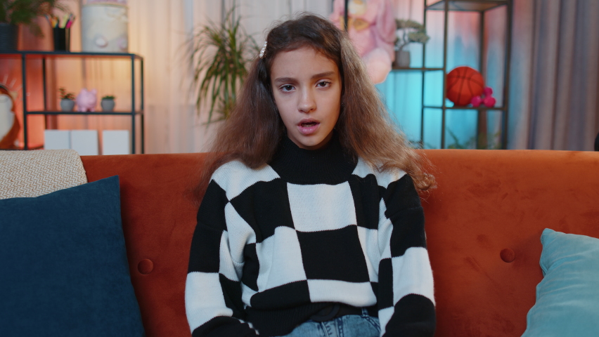 Dislike! Upset girl showing thumbs down sign gesture, expressing discontent, disapproval, dissatisfied bad work at modern home apartment indoors. Displeased young child, kid in living room on sofa
