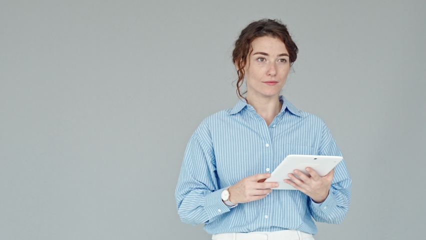 Young professional business woman leader, coach, manager speaker holding tablet, talking making presentation, communicating with audience during business training standing at grey wall.