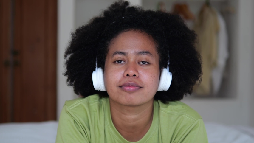 A young afro woman with black beautiful curly hair with a funny, calm and friendly mood spends her free leisure time lying down, listening to music wearing headphones on the bed a cozy room.