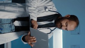 Vertical video: Portrait of smiling doctor wearing medical coat and stethoscope holding laptop computer, typing health care expertise. Clinical staff working over hours in hospital office. Medicine