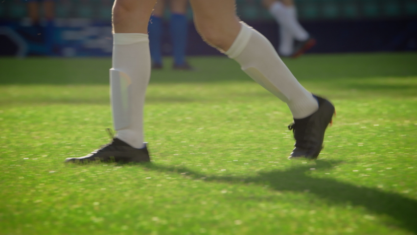 Focus on Legs of a Professional Soccer Player Walking on the Green Grass Stadium Field. Players Resting Between Attack and Defense. Low Angle Ground Shot.