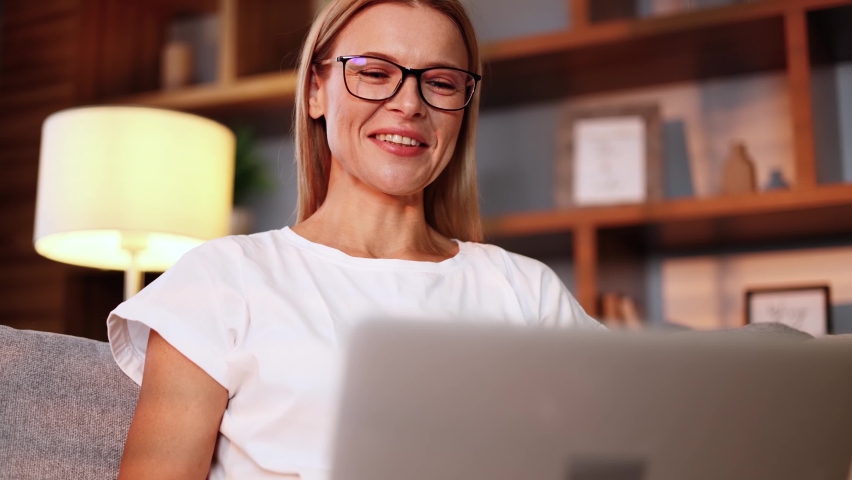 Portrait of charming middle aged woman with eyeglasses laying on the sofa in cozy living room and talking on laptop by video chat conference meeting. Remote work freelance or distance relashionship.