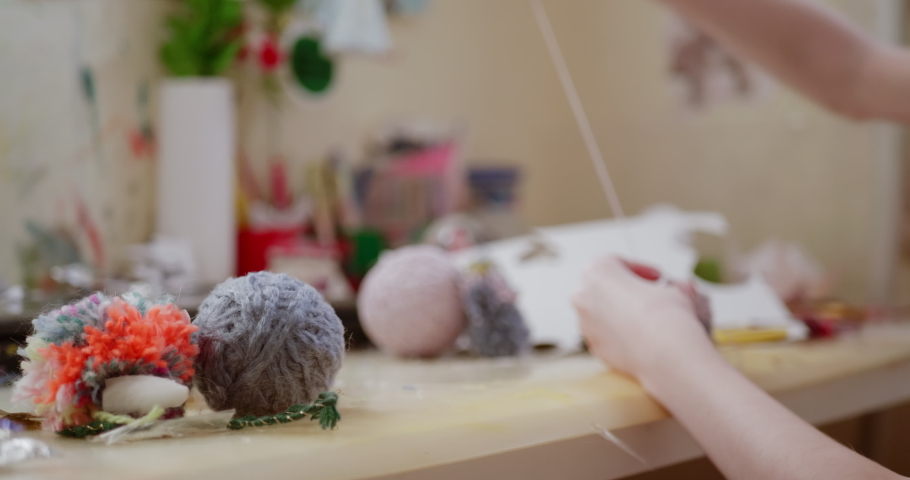 Close-up on blury child hands making pompom crafts with light coloured yarn at table, handheld shot and selective focus, copy space.