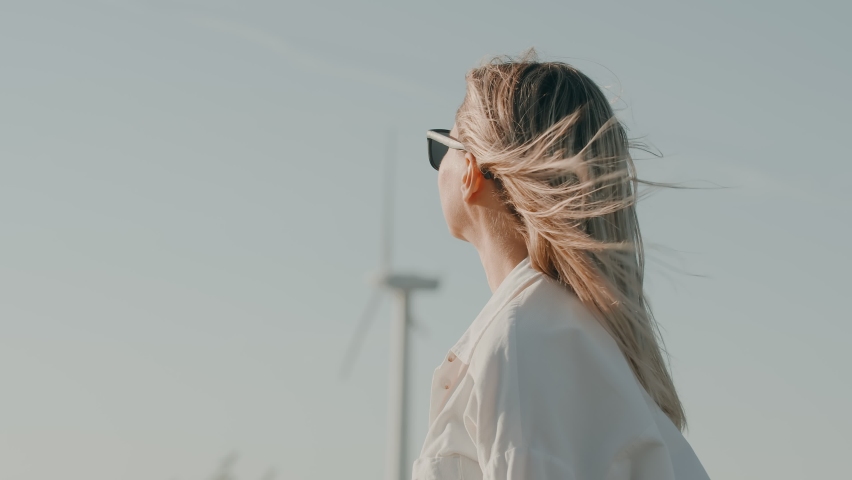 Young beautiful caucasian woman standing outdoor with swaying on the wind blonde long hair with working wind turbines in the background. Modern woman thinking about her future and environment Royalty-Free Stock Footage #1097305907