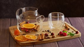 Making fruit tea, pouring hot water in transparent glass teapot filled with orange fruit, cinnamon sticks and berries, cup placed on wooden table with mint and raspberry, slow motion 4k video footage