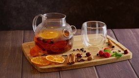 Woman making fruit tea with orange slices, cranberries, cinnamon sticks, adding ingredients into glass teapot placed on table with cup, hot vitamin winter drink, preparing spice tea, 4k video clip