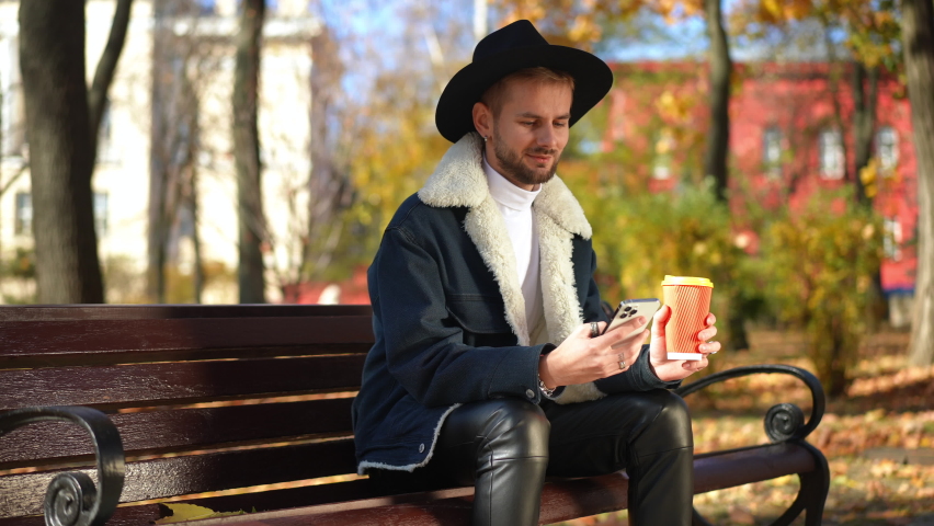 Smiling LGBT man drinking coffee messaging in smartphone app sitting on bench in sunny park. Positive confident handsome guy enjoying leisure outdoors on autumn day Royalty-Free Stock Footage #1097317425