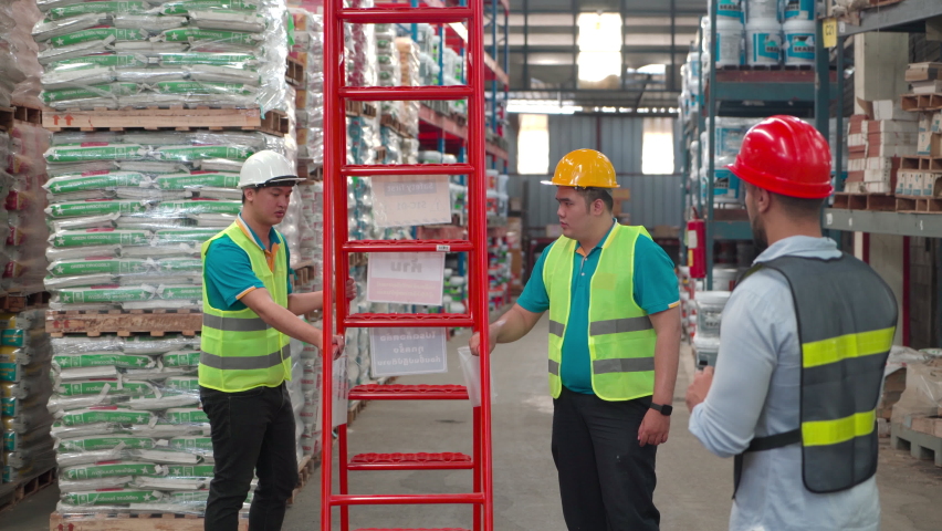Caucasian male engineer guides two male workers is draging red ladder. Employees wear safety hat and reflective vests working in a warehouse, team work, helping each other.