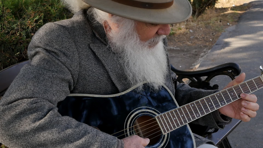 An elderly stylish man with a gray beard plays an acoustic guitar while sitting on a bench outdoors. Senior man guitarist wears a hat and gray coat and wears a nice fluffy beard like a movie wizard