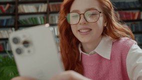 Portrait of happy young student chatting by mobile phone in university library. Attractive Woman with Red Hair and Eyeglasses using social media application browsing on smartphone. Library Background