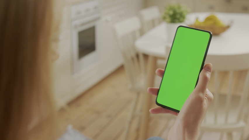 Handheld Camera: Back View of Young Woman at Home Kitchen Sitting on a Chair using With Green Mock-up Screen Smartphone. Girl is Surfing Content With Touching Tapping on Center Screen