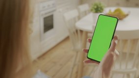 Handheld Camera: Back View of Young Woman at Home Kitchen Sitting on a Chair using With Green Mock-up Screen Smartphone. Girl is Surfing Content With Touching Tapping on Center Screen - Powered by Shutterstock - Get 15% off with code: PIKWIZARD15