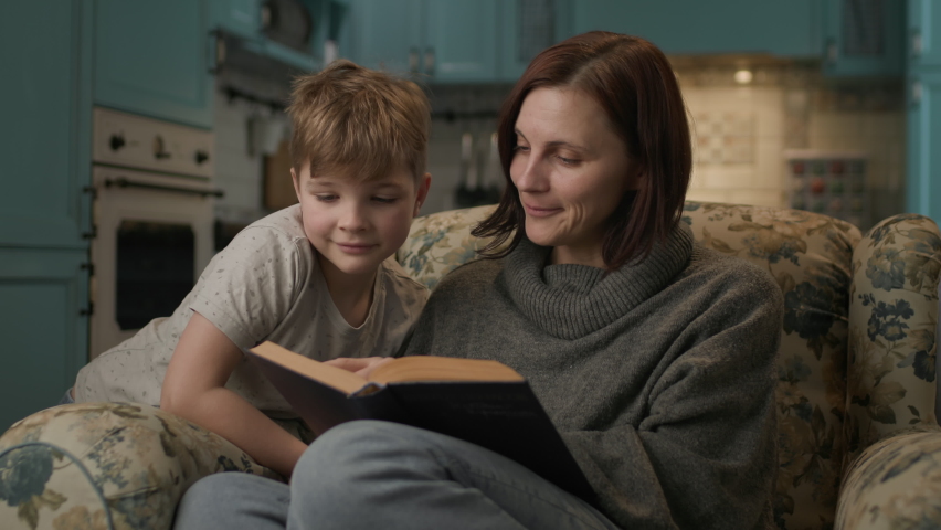 Mom reads paper book to her son. 30s woman enjoys reading literature with her kid sitting on cozy couch at home. | Shutterstock HD Video #1097328553