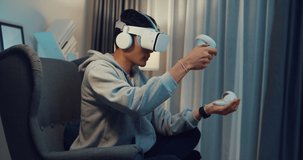 Young Asian man gamer wear gray hoodie with VR controllers headset hold joystick controller creating 3D art sit on sofa living room home at night. Futuristic artwork virtual reality concept.