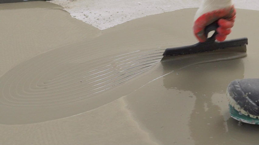 Repairman in protective gloves and uniform using metal spatula for aligning liquid self-levelling floor screed. Worker spreading liquid cement solution on construction site. Royalty-Free Stock Footage #1097330469