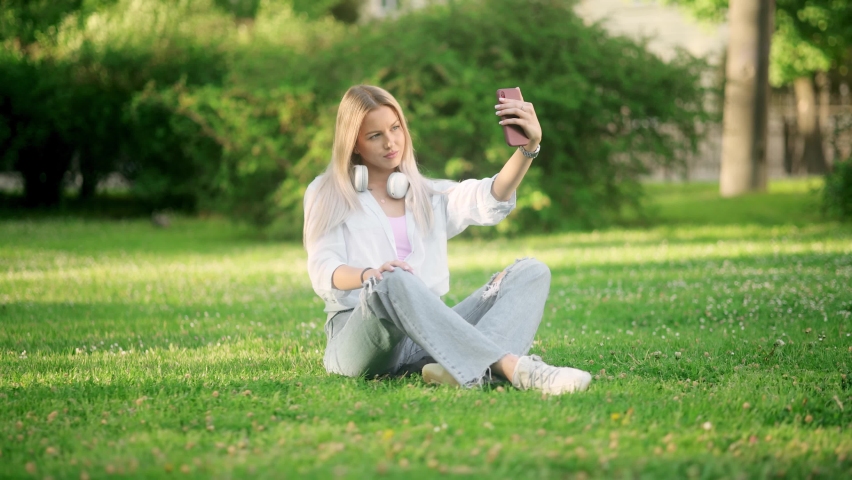 Young pretty blonde woman in jeans and a white shirt sits on the grass in the park and takes a selfie | Shutterstock HD Video #1097338757