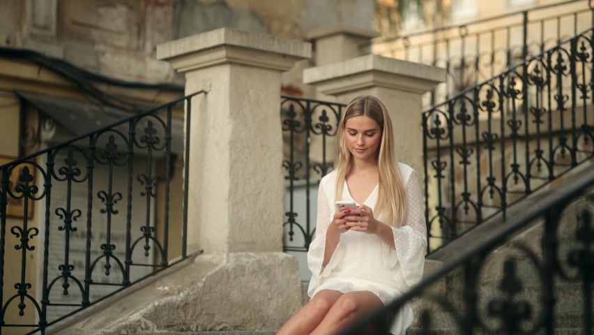 Portrait smiling young woman in a white dress with blonde hair sitting on stairs using mobile phone | Shutterstock HD Video #1097339683