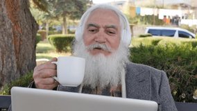 Stylish intelligent senior man holding a mug with a hot drink is talking on a video link using a laptop and white wireless headphones. Modern advanced grandfather calls relatives by videoconference