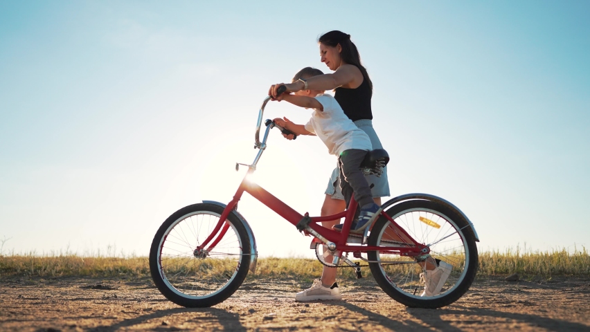 Happy family concept. Mom teaches her son to ride bike on green grass. Child rides bicycle along rural road. Green energy. Mom teaches her son to ride bike for the first time in park at sunset. Royalty-Free Stock Footage #1097349499