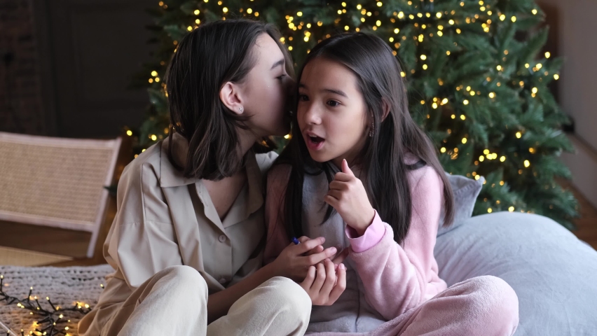 Two cute sisters, friends share the news, whisper secrets in your ear, sitting on the bed against the backdrop of the Christmas tree. Cute children celebrate winter holidays. Festive mood. | Shutterstock HD Video #1097350099