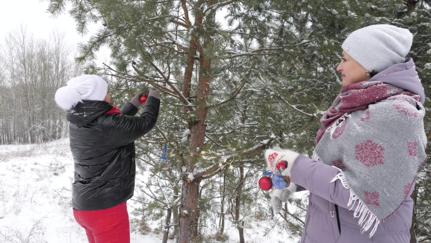 Two adult women in warm clothes decorate fir tree with New Year's balls in winter forest | Shutterstock HD Video #1097350383
