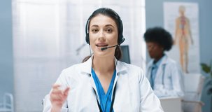 Close up portrait of Caucasian pretty young woman doctor in white medical coat waving hand looking at camera and speaking in headset sitting in hospital cabinet. Healthcare, video consultation