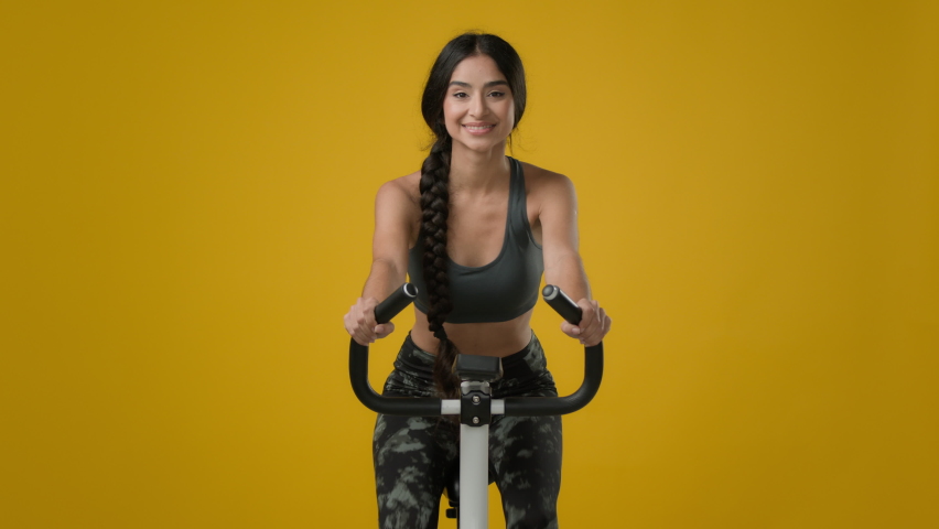 Indian slim happy smiling sporty woman female lady athletic girl cyclist biking flexing cycling riding on exercise bike sport equipment fitness workout cardio ride in yellow studio thumb up gesture Royalty-Free Stock Footage #1097360667
