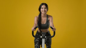 Indian slim happy smiling sporty woman female lady athletic girl cyclist biking flexing cycling riding on exercise bike sport equipment fitness workout cardio ride in yellow studio thumb up gesture - Powered by Shutterstock - Get 15% off with code: PIKWIZARD15